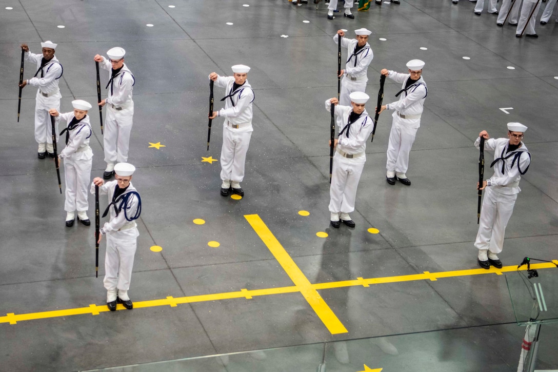 A group of sailors stand in formation, each holding a staff with both hands to one side of their bodies, as other sailors stand behind them.