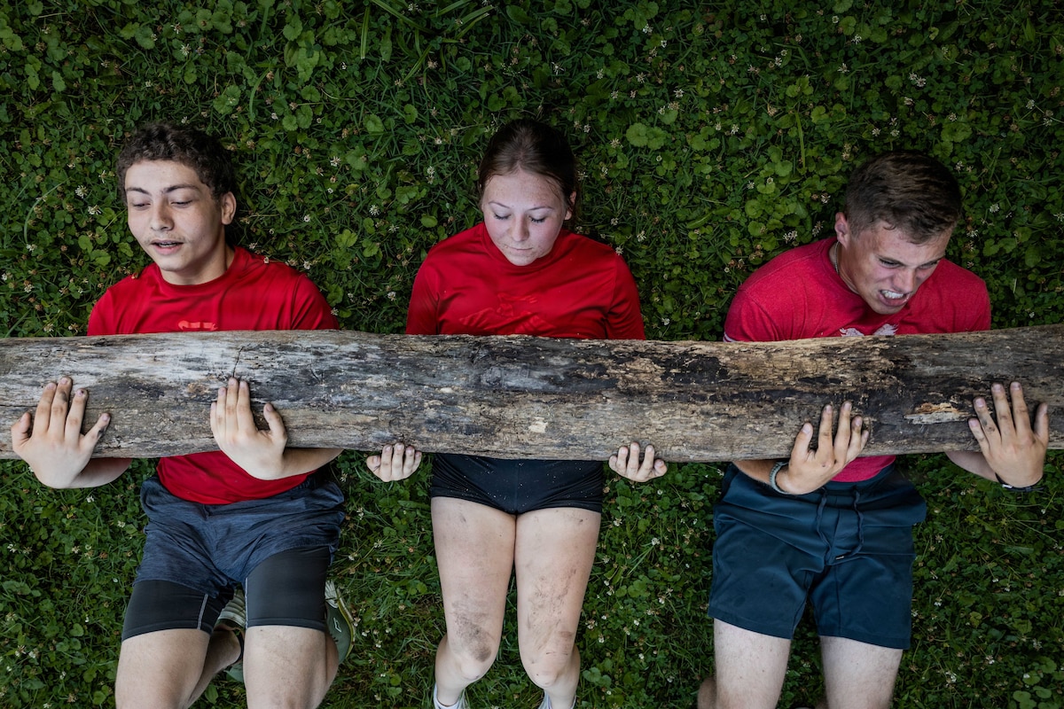 Three people in red T-shirts do crunches while holding a log. Two of the people have slight smiles on their faces.