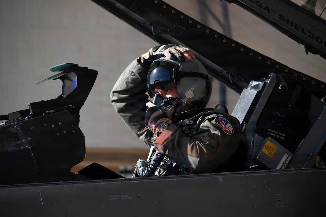 A pilot seated in a fighter jet secures an oxygen mask with one hand, while holding the top of their helmet with the other hand.