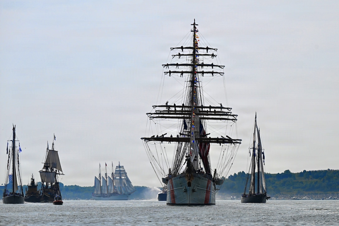 A sailing ship with tall masts sails through a body of water with other sailing vessels and boats behind it on a gray, overcast day.