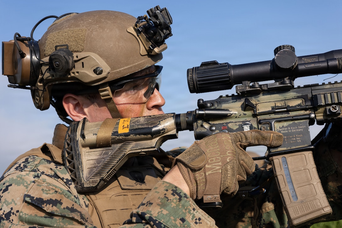 A Marine wearing a camouflage uniform and helmet looks through a scope mounted on a military rifle outdoors on a bright, sunny day.