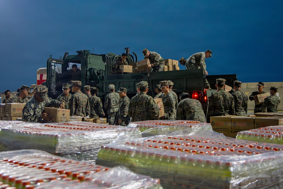 A group of approximately two dozen Marines in camouflage uniforms remove boxes of supplies from the back of a truck during low-light conditions.