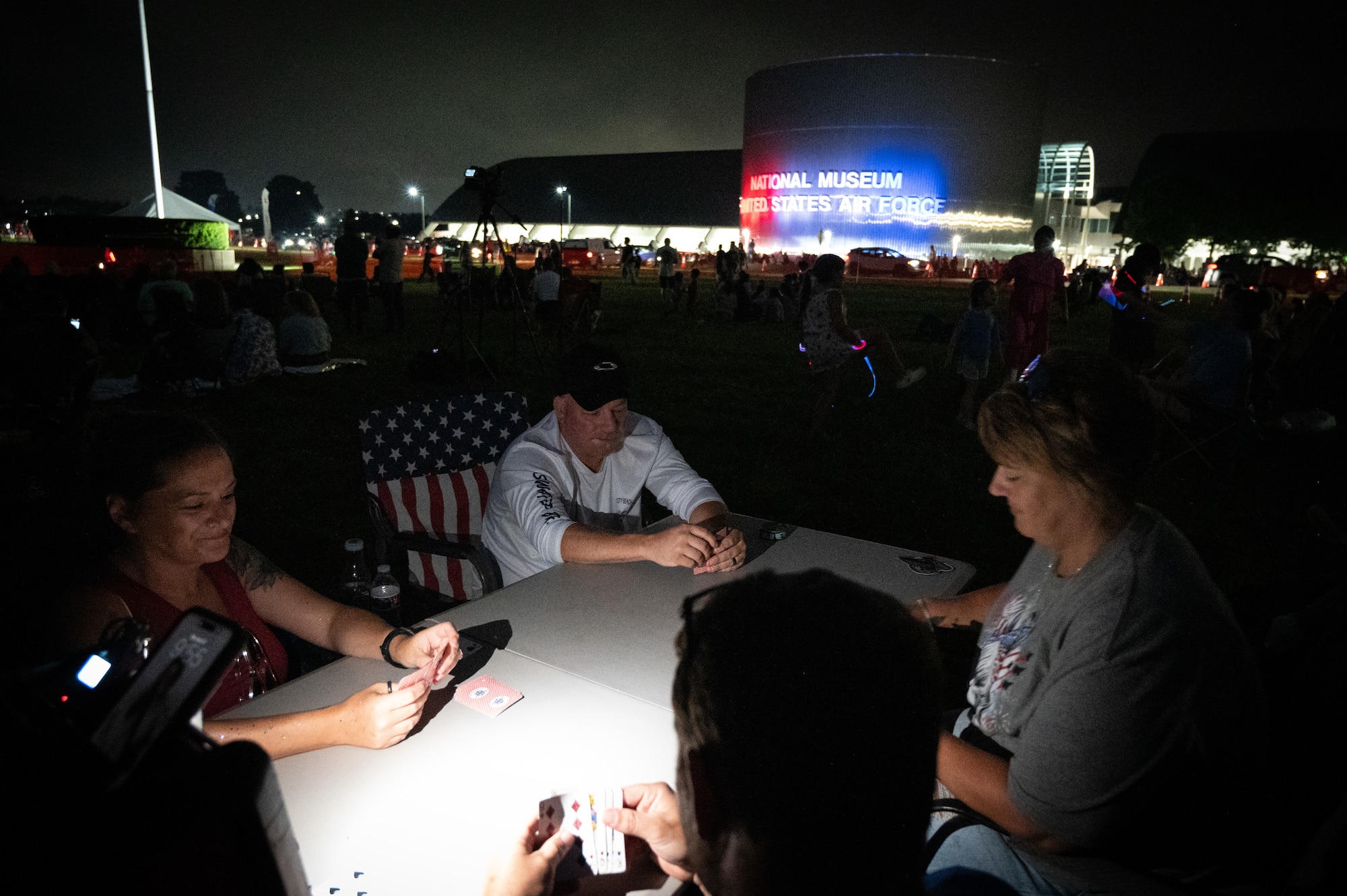 Family sitting around a fold-up table in lawn chairs playing cards by phone flashlight in front of the museum.