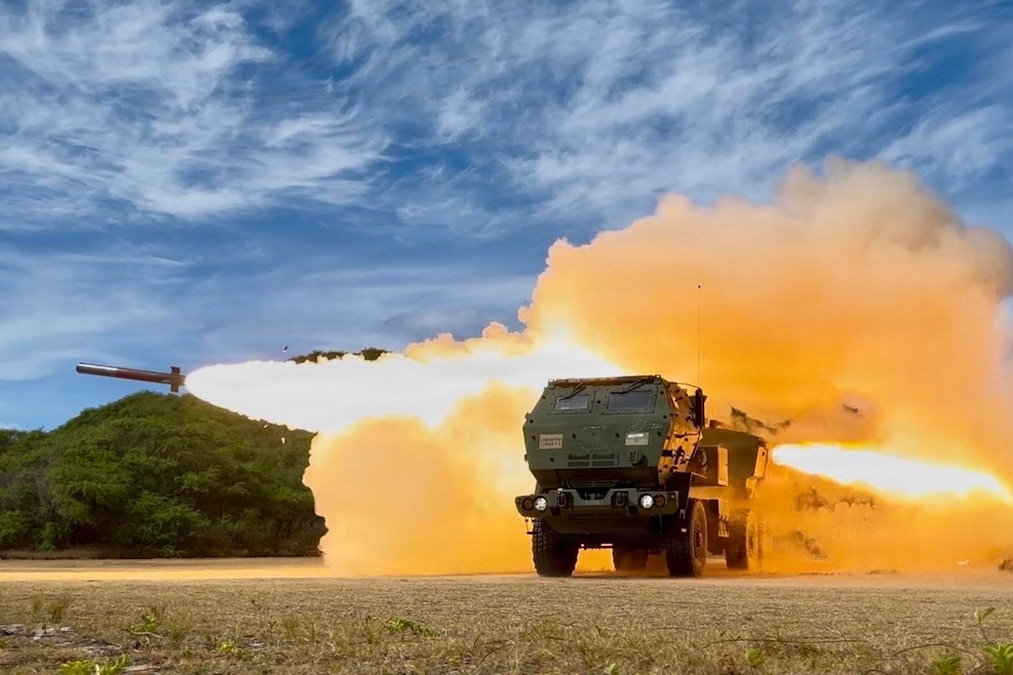A rocket flies from a truck-mounted launcher amid a large ball of flames outdoors during daytime with foliage in the background.