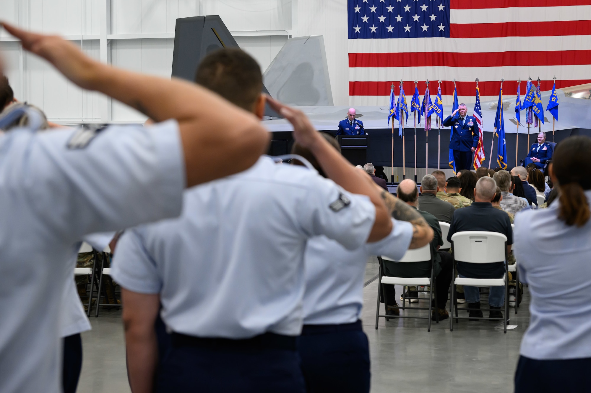 A wide-angle photo taken from the back of a military change of command ceremony. In the foreground, the backs of Airmen standing in formation are visible as they render a hand salute. In the middle ground, an seated audience of civilian and military guests separates the formation from the stage. In the background on the stage, Air Force Colonel Eric B. Quidley stands saluting back, flanked by organizational military flags.
