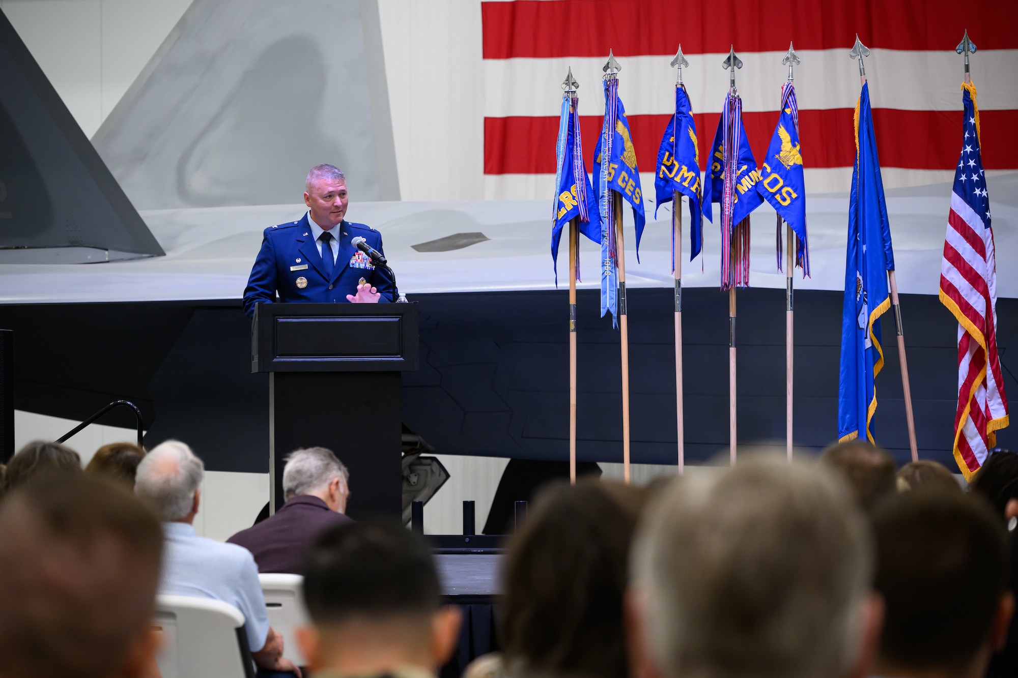 Air Force Colonel Eric B. Quidley stands at a wooden podium speaking into a microphone during an indoor change of command ceremony. He is wearing a formal blue service dress uniform. The United States flag and the 75th Air Base Wing organizational flag stand behind him on the stage.