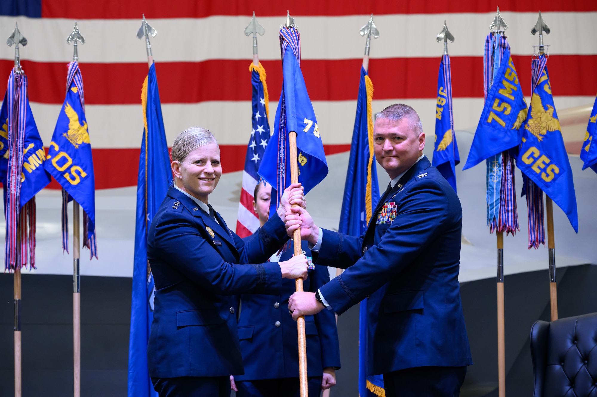 A formal military change of command ceremony on a stage. Air Force Lieutenant General Jennifer Hammerstedt, in service dress uniform, is passing a military guidon flag to Colonel Eric B. Quidley, who is standing on the right. A third service member in uniform stands at attention in the background. Official organizational flags and the American flag are displayed behind them.