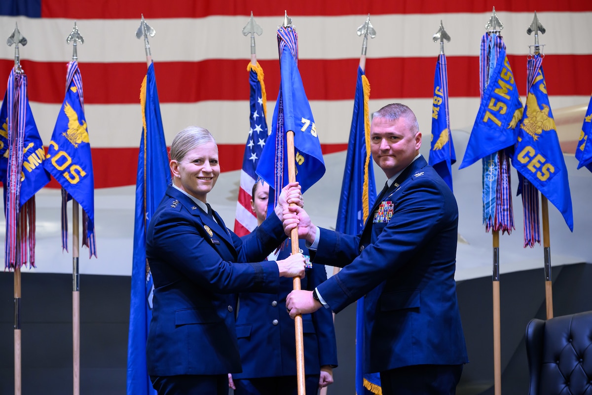 A formal military change of command ceremony on a stage. Air Force Lieutenant General Jennifer Hammerstedt, in service dress uniform, is passing a military guidon flag to Colonel Eric B. Quidley, who is standing on the right. A third service member in uniform stands at attention in the background. Official organizational flags and the American flag are displayed behind them.