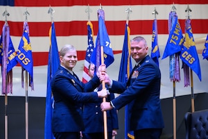A formal military change of command ceremony on a stage. Air Force Lieutenant General Jennifer Hammerstedt, in service dress uniform, is passing a military guidon flag to Colonel Eric B. Quidley, who is standing on the right. A third service member in uniform stands at attention in the background. Official organizational flags and the American flag are displayed behind them.
