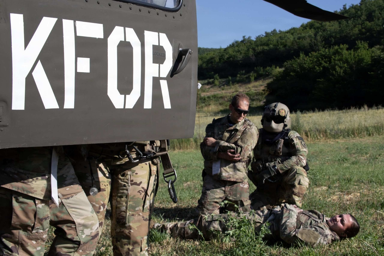 Two people in camouflage military uniforms tend to another person in similar attire lying on the ground in a field.