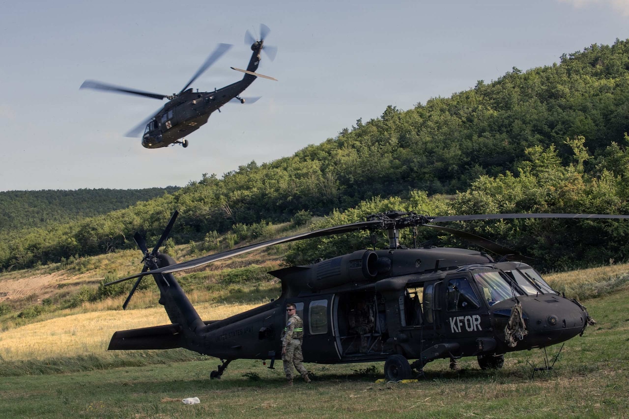 A military helicopter takes off from a field while another helicopter is still on the ground.