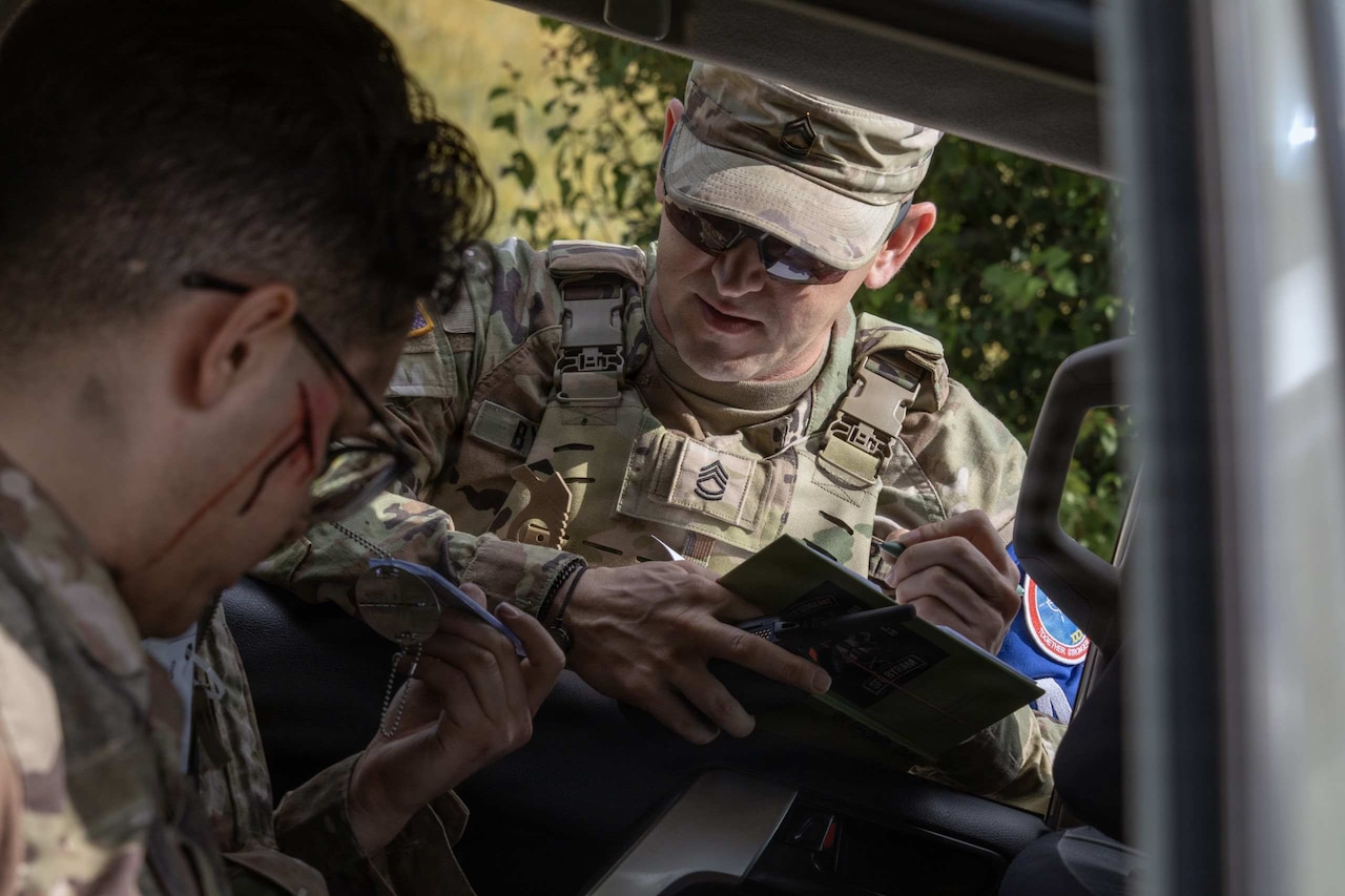 A man in a camouflage military uniform writes in a notebook while talking to another man in similar attire sitting in car.