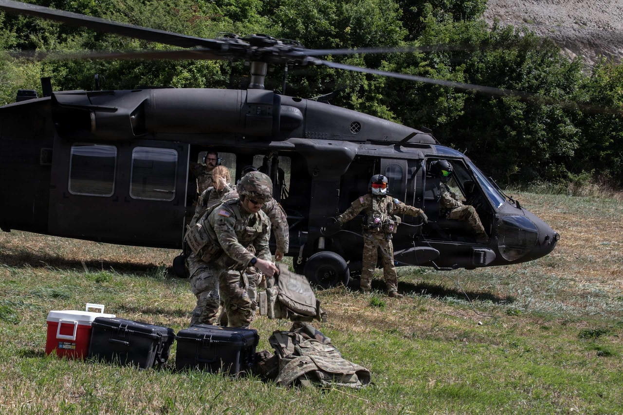A man in a camouflage military uniform grabs gear from the ground in an open field as another person in similar attire takes gear to a helicopter sitting in the field in the background.