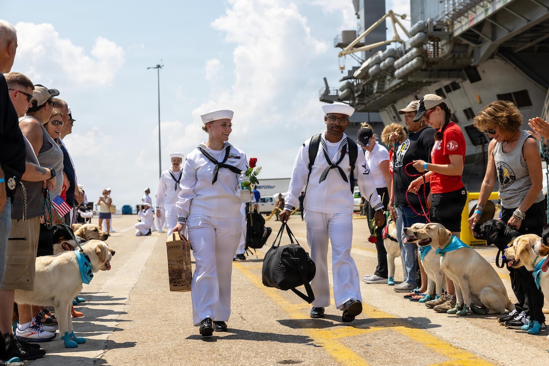 Sailors in white uniforms carry luggage while walking in between a parallel line of people holding Labrador retrievers with blue bandannas on their necks.