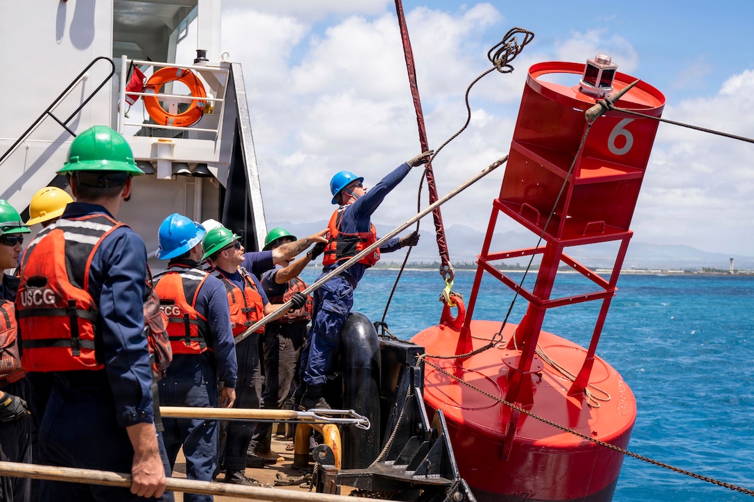 Coast Guardsmen in life vests and hard hats aboard a ship at sea use a line to keep an orange buoy in place as a fellow Coast Guardsman throws a rope on the top.
