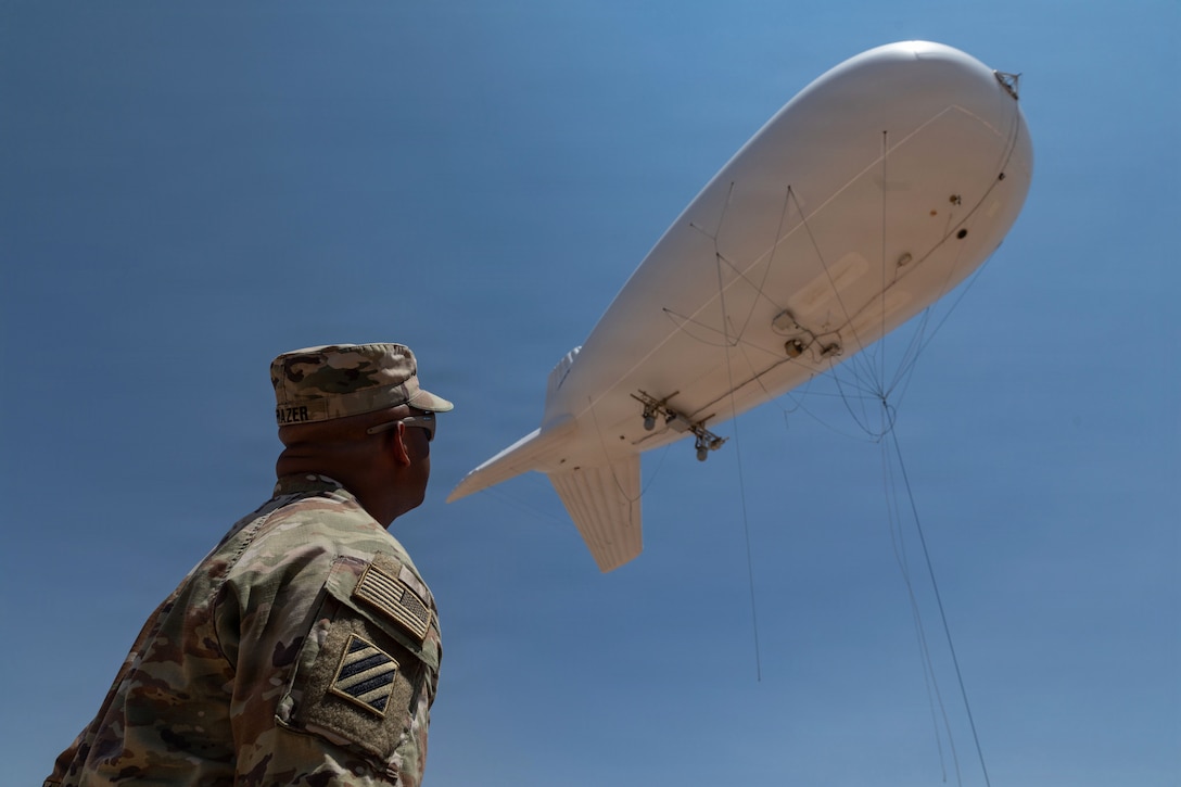 A soldier looks up at a white, tethered surveillance system as it moves through a blue sky.