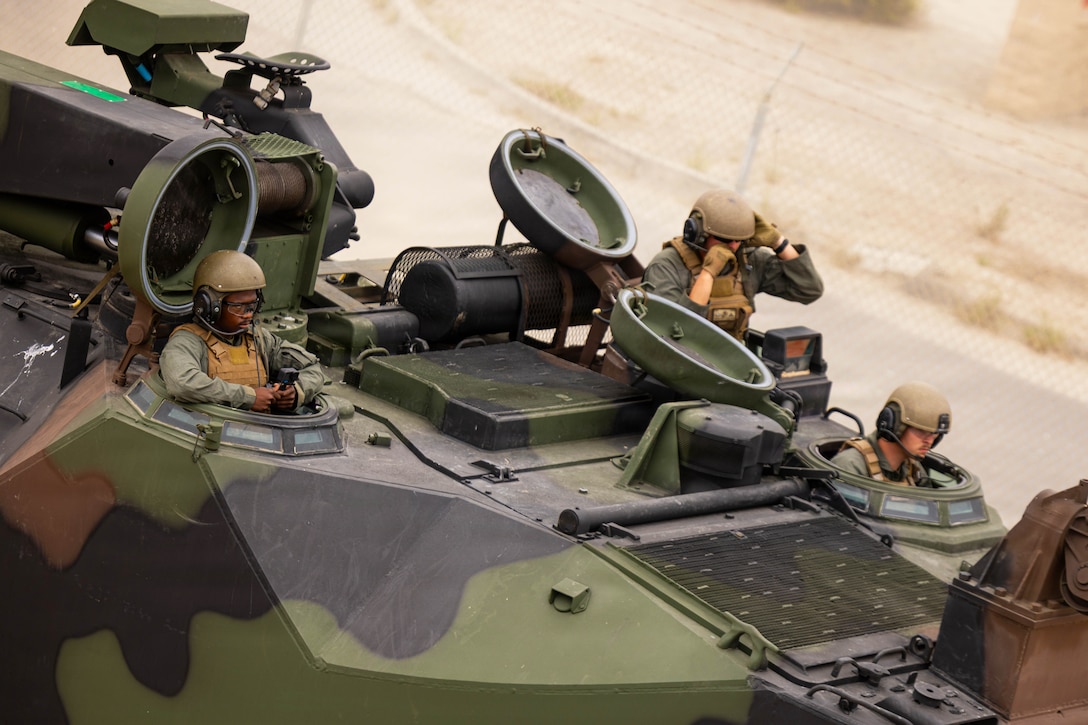 Three Marines look out of open hatches at the top of an amphibious assault vehicle. A fence and dirt are in the background.