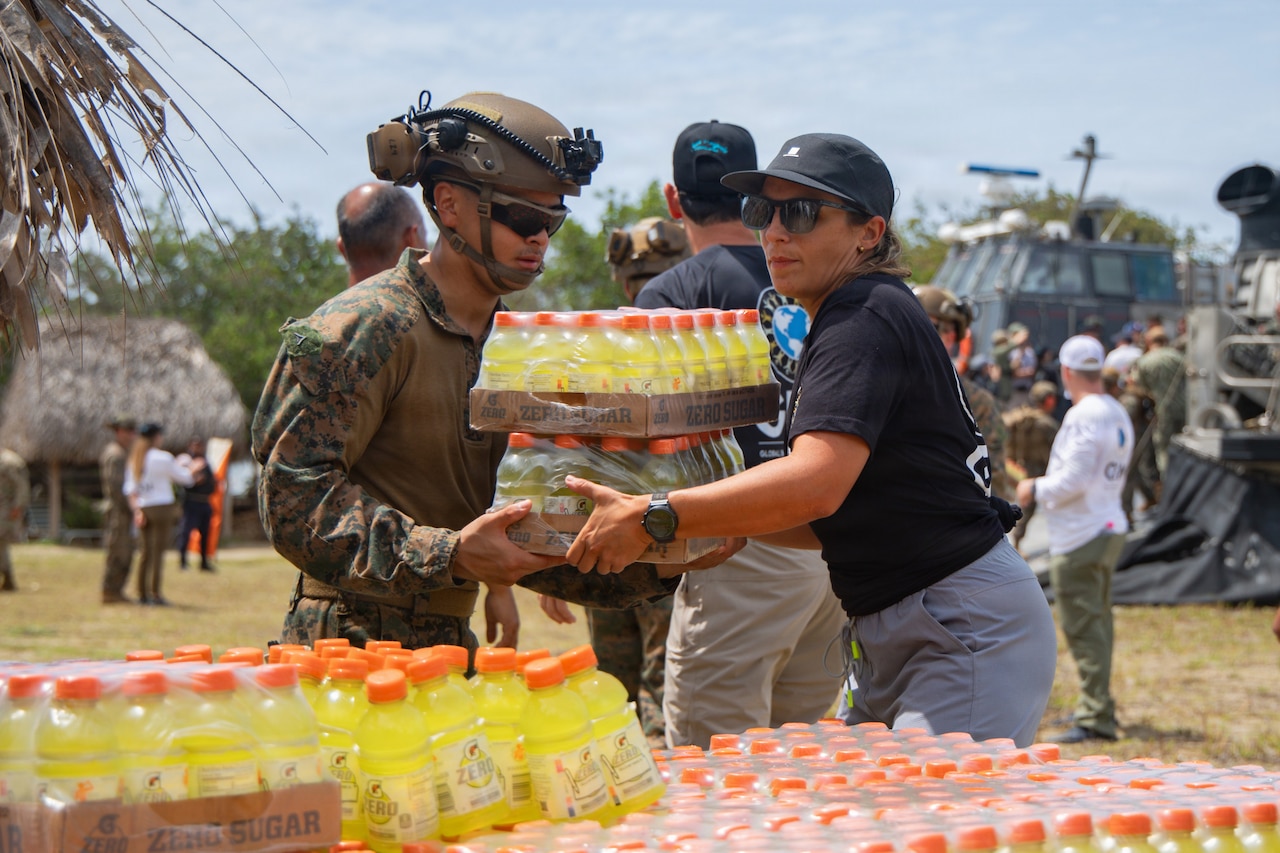 A Marine hands someone cases of yellow Gatorade as other orange-topped Gatorade bottles sit on a table next to them. Trees and other people are in the background.