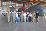 A group of people, some wearing military uniforms, stand in front of an aircraft in a hangar.