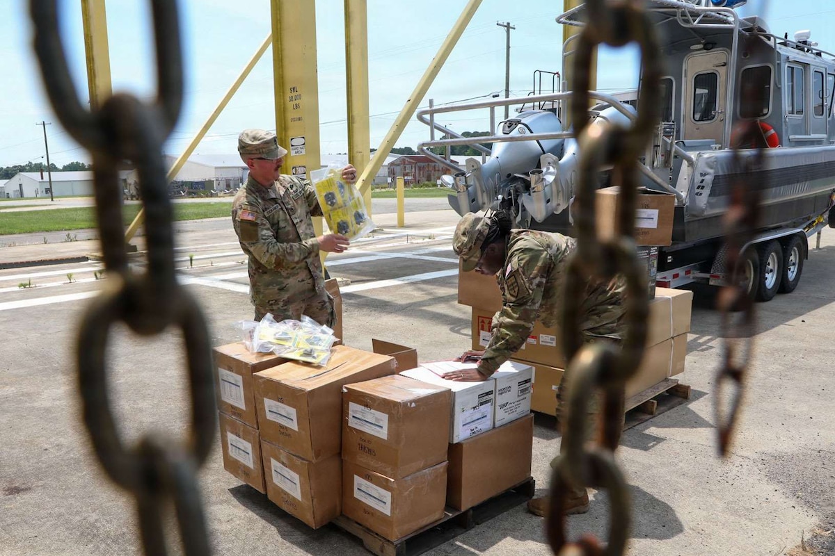 Two people wearing camouflage military uniforms inspect a pallet of packages while standing outside; there is a boat on a trailer in the background.