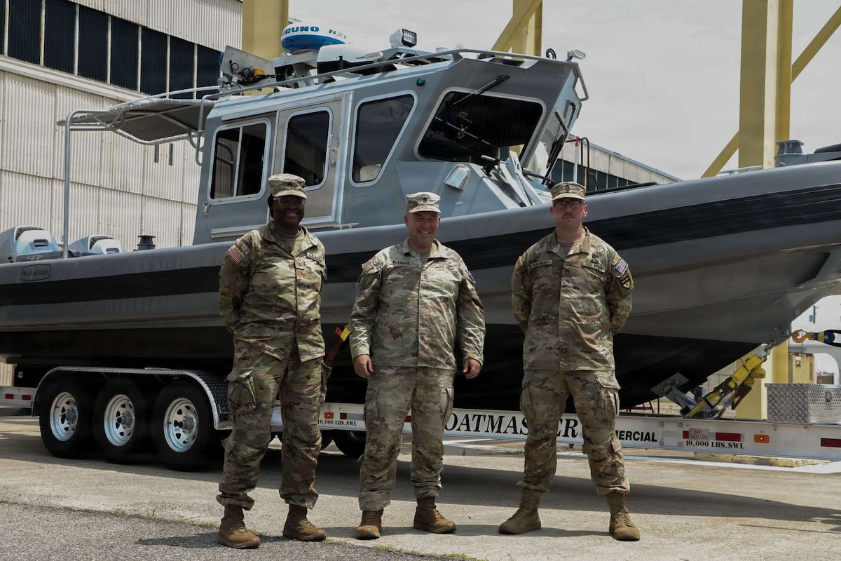Three people wearing camouflage military uniforms pose for a photo while standing in front of a boat on a trailer.