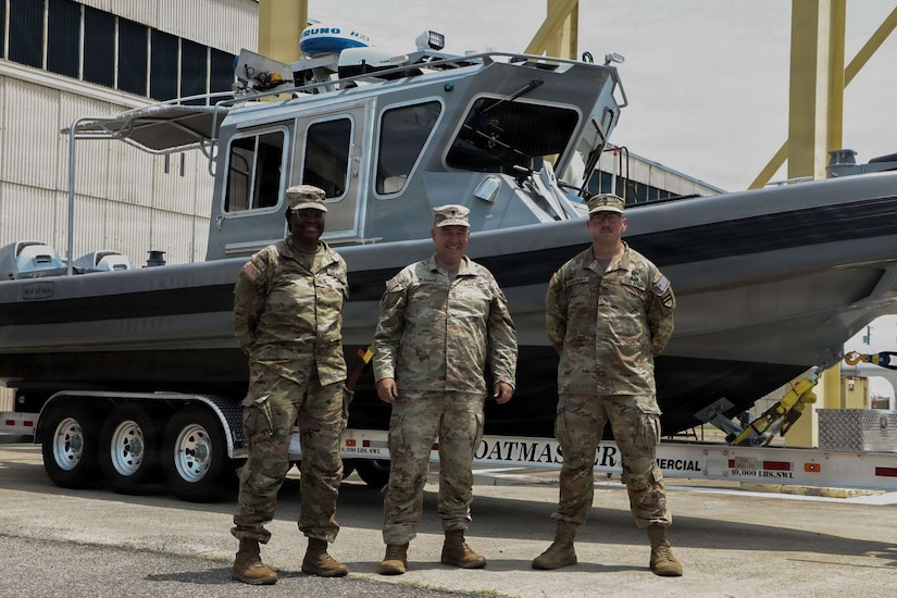 Three people wearing camouflage military uniforms pose for a photo while standing in front of a boat on a trailer.