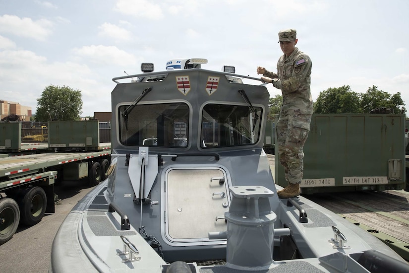 A man wearing a camouflage military uniform walks around the edge of a boat sitting on a trailer.