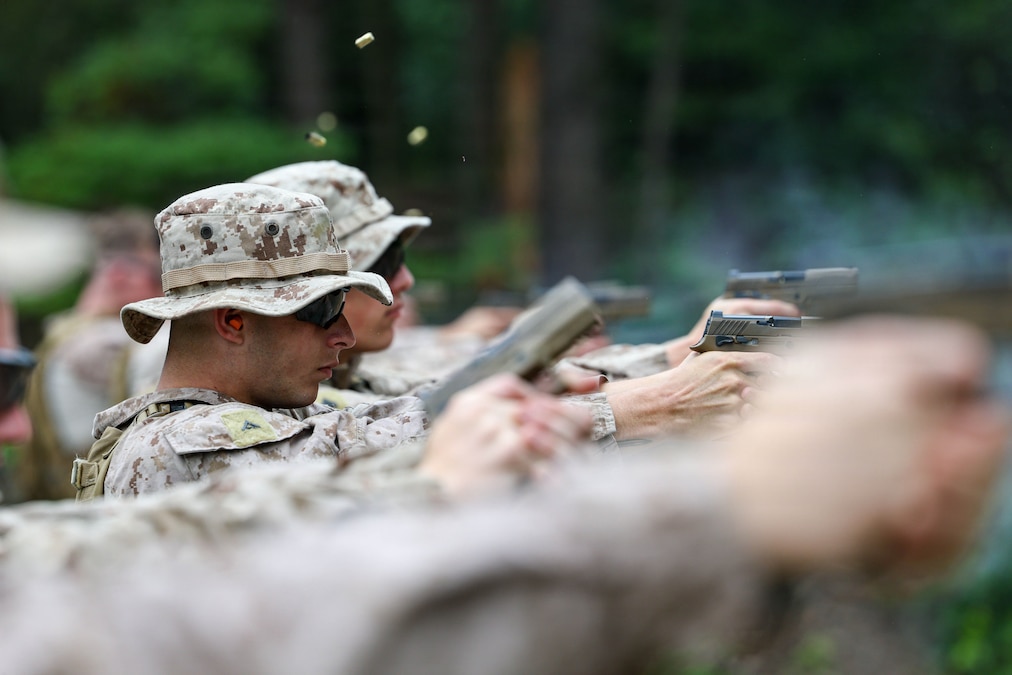 A blurred arm holding a pistol is seen in front of a row of soldiers firing pistols. A few shell casings are in the air.