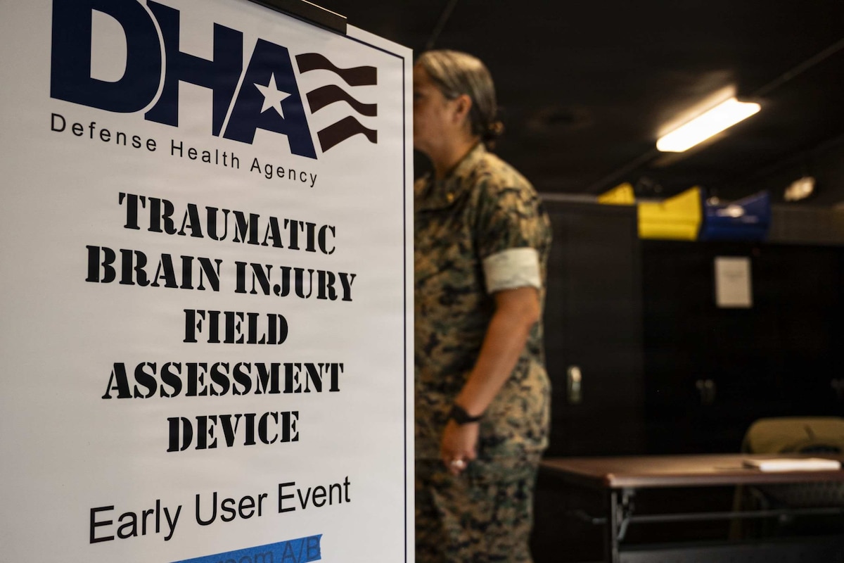 A sign for a traumatic brain injury training event stands in a small room as a woman wearing a camouflage military uniform walks by.