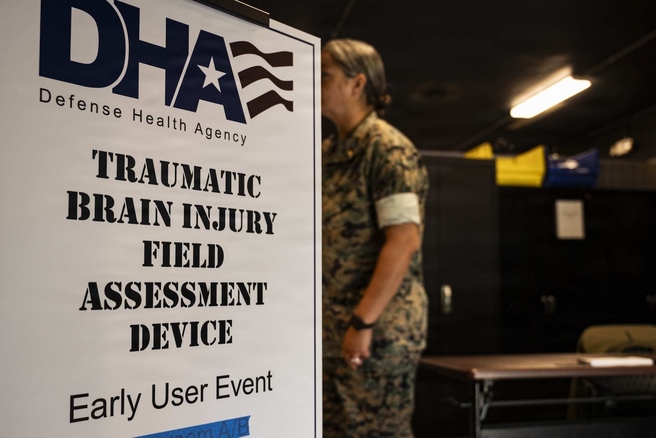 A sign for a traumatic brain injury training event stands in a small room as a woman wearing a camouflage military uniform walks by.