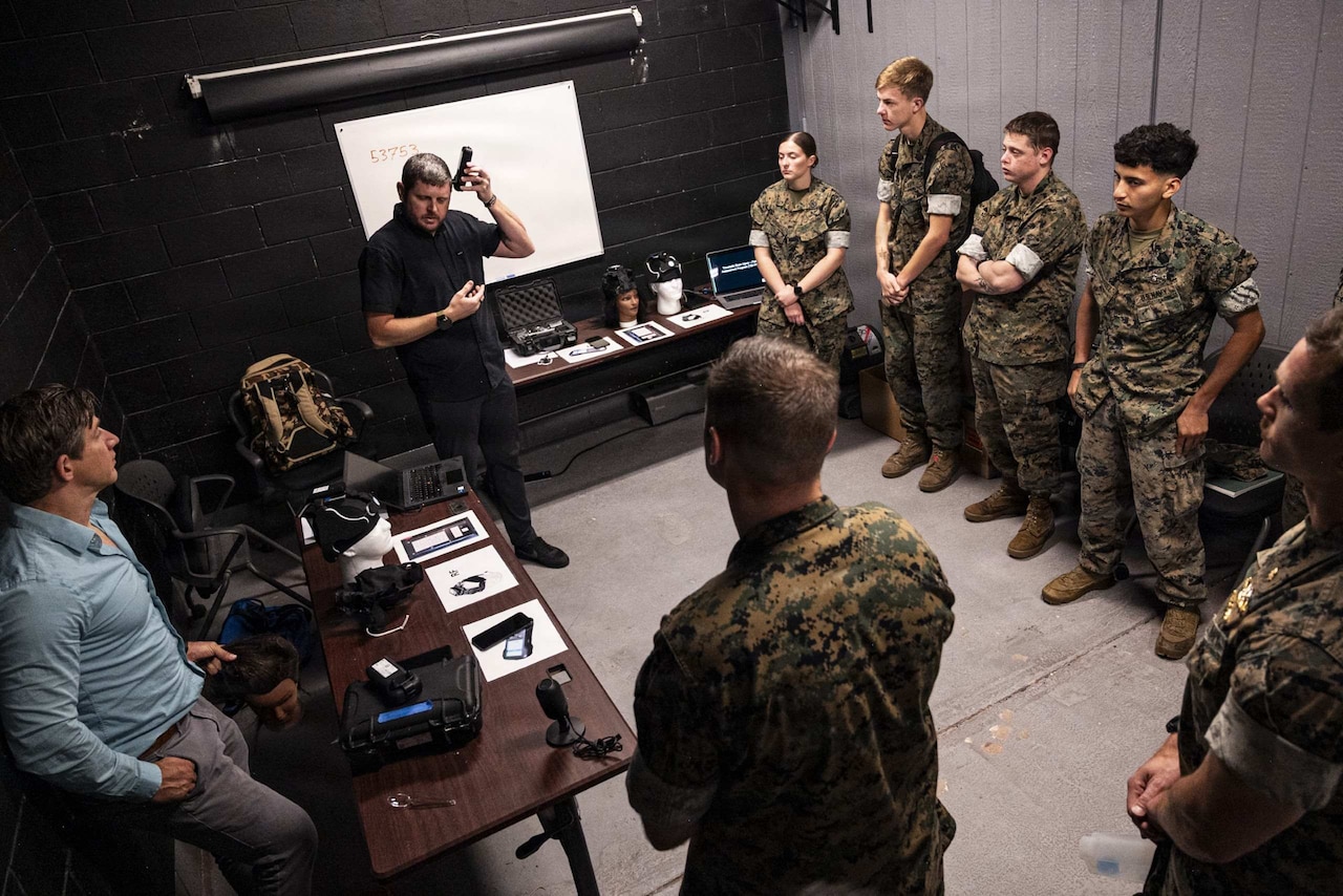 A man wearing a black shirt stands near a white board in front of a room with multiple people wearing camouflage military uniforms.