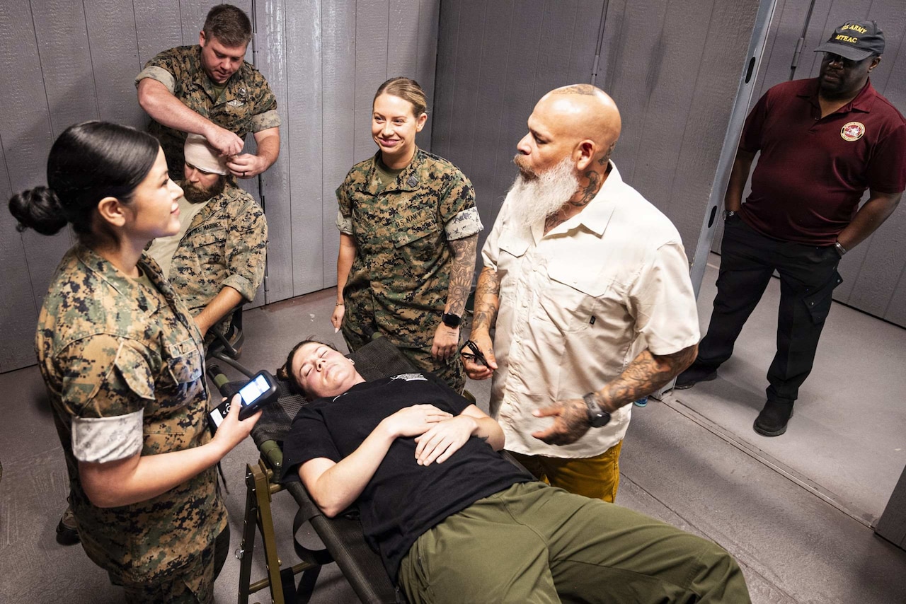A person lies on a cot as other people, some wearing camouflage military uniforms, stand and speak to each other.