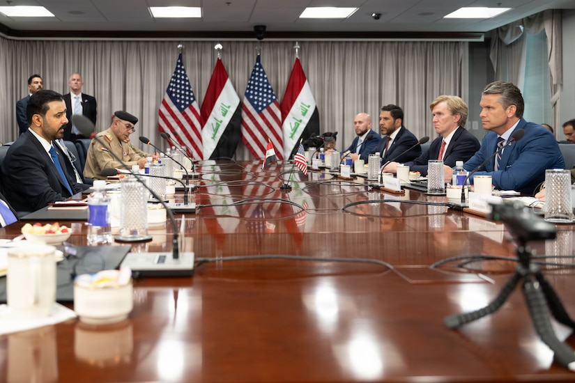 Several men in suits and military uniforms talk while sitting at a table as the American and Iraqi flags are displayed in the background.