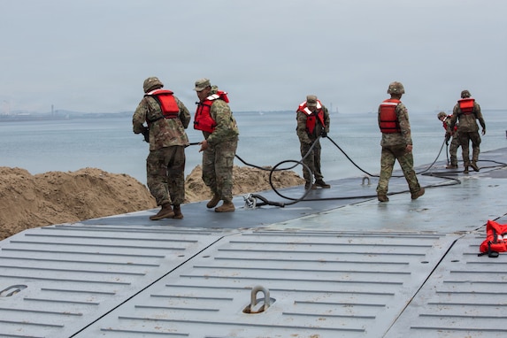 Several U.S. Army soldiers wearing combat helmets and safety vests stand in the shallow water and surf of Dogu Beach, physically pulling heavy mooring lines to secure a large, flat, modular metal Trident Pier structure extending into the ocean.