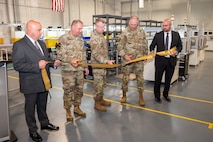Five men cutting a golden ceremonial ribbon with large gold scissors. Three center men are in Army Uniform, both men on their sides are wearing suits.