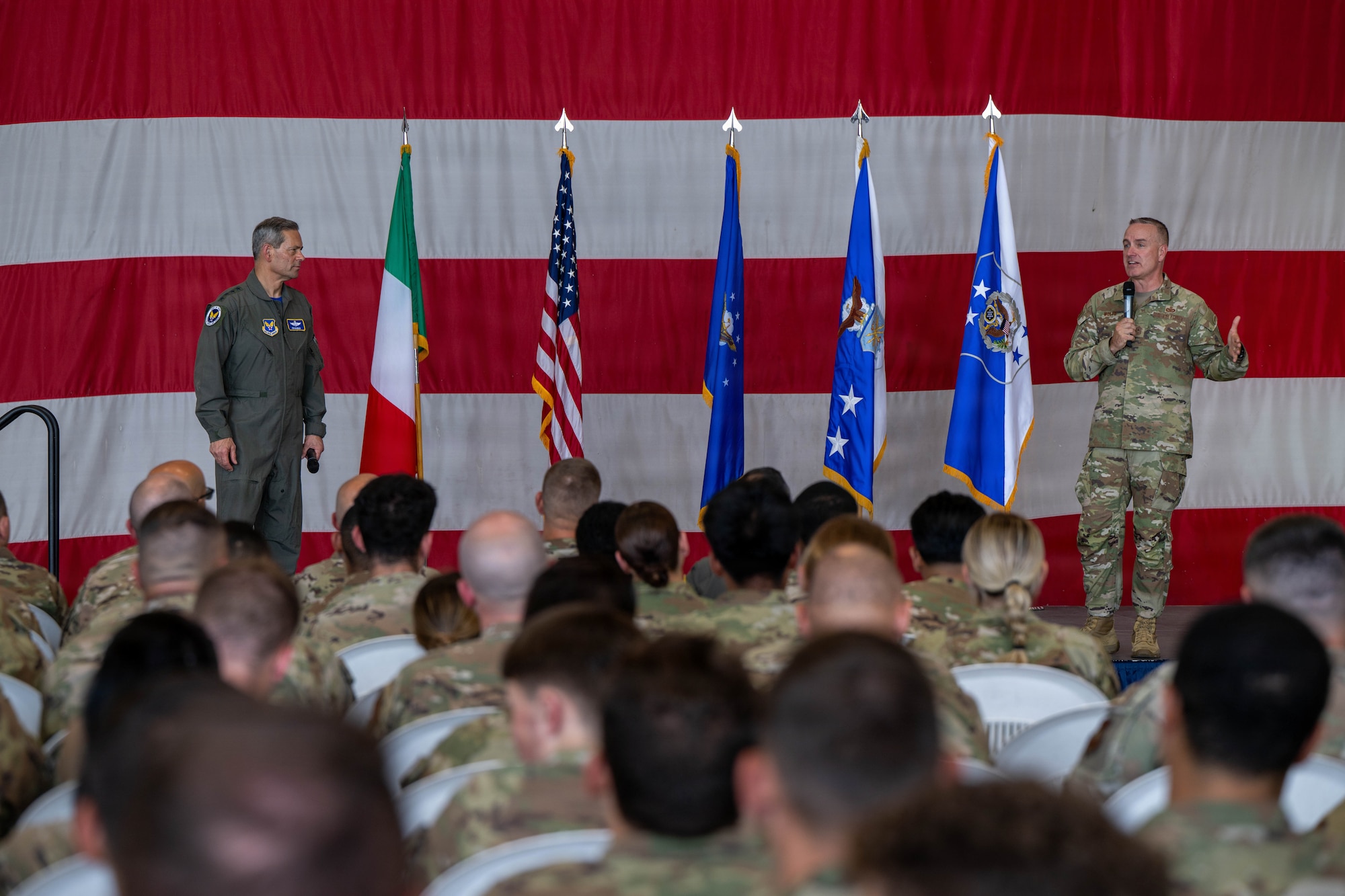 Two men stand on a stage speaking to a large audience.
