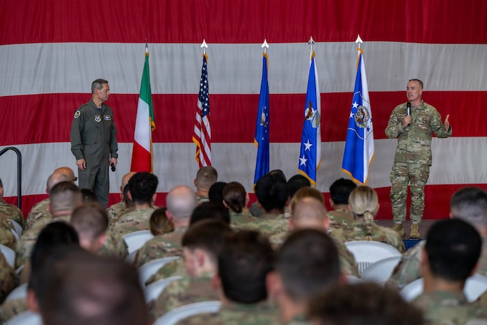 Two men stand on a stage speaking to a large audience.