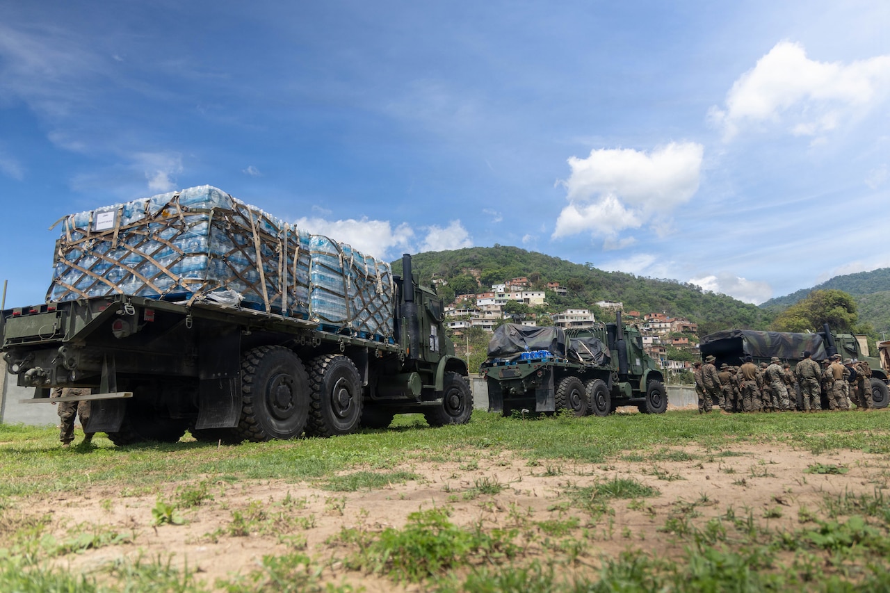 Military trucks loaded with pallets of drinking water and other supplies are parked on a grassy field with hills in the background on a sunny day.