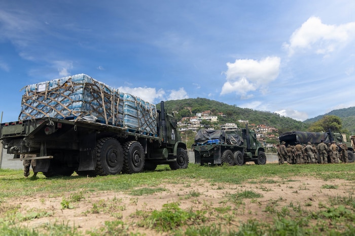 Military trucks loaded with pallets of drinking water and other supplies are parked on a grassy field with hills in the background on a sunny day.