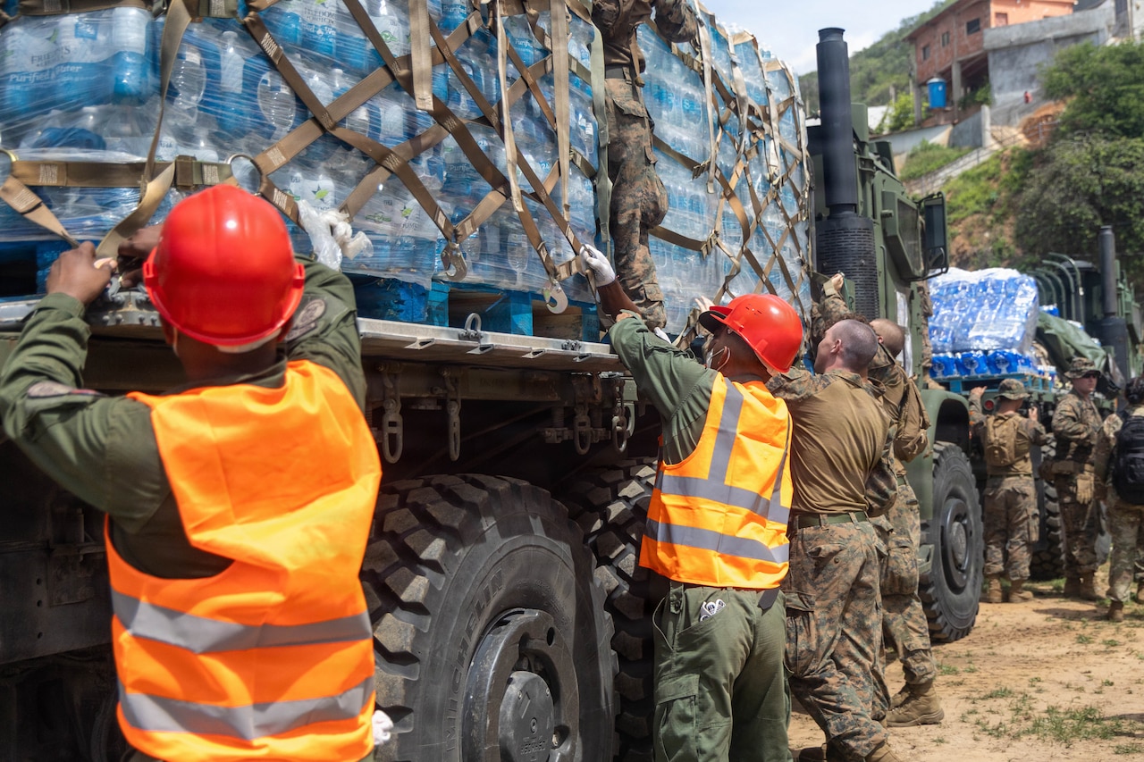 People wearing camouflage and olive drab military uniforms, some of whom are also wearing orange safety vests and red helmets, work to remove pallets with bottled drinking water from military trucks during daytime.