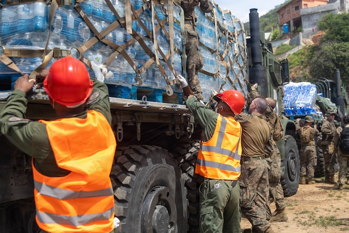 People wearing camouflage and olive drab military uniforms, some of whom are also wearing orange safety vests and red helmets, work to remove pallets with bottled drinking water from military trucks during daytime.