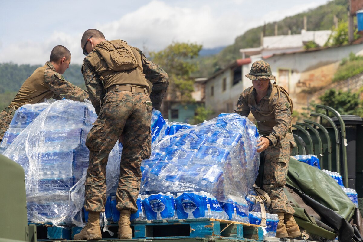 Three Marines in camouflage uniforms remove pallets of water from the back of a military truck.