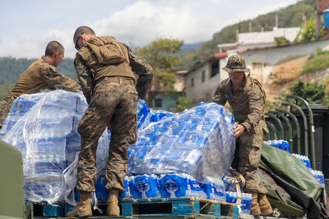 Three Marines in camouflage uniforms remove pallets of water from the back of a military truck.