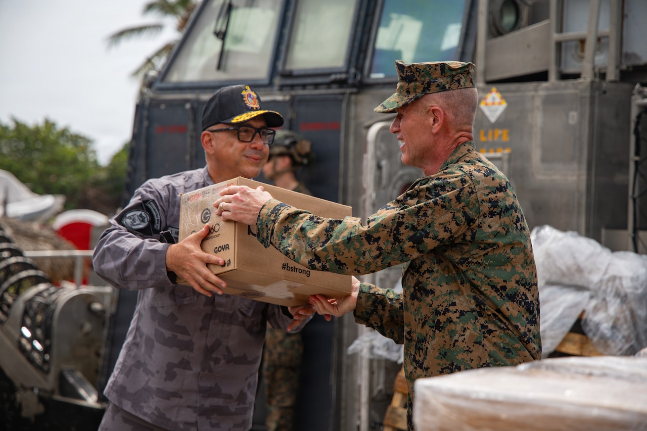 A Marine wearing a camouflage uniform hands a cardboard box to a military service member wearing a different color camouflage uniform outdoors during daylight.