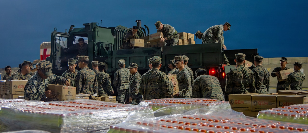A group of about two dozen Marines in camouflage uniforms remove boxes of supplies from the back of a military truck under low-light conditions.