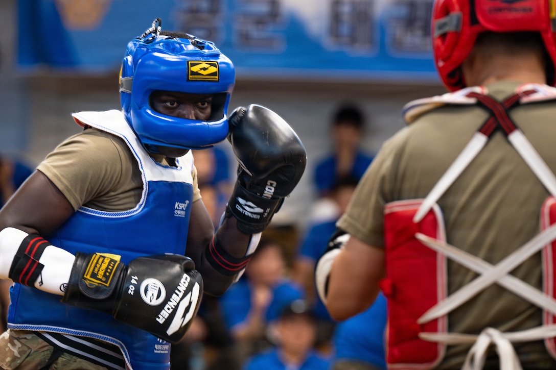 Two service members in red and blue boxing gear face off as seated audience members watch from the blurred background.