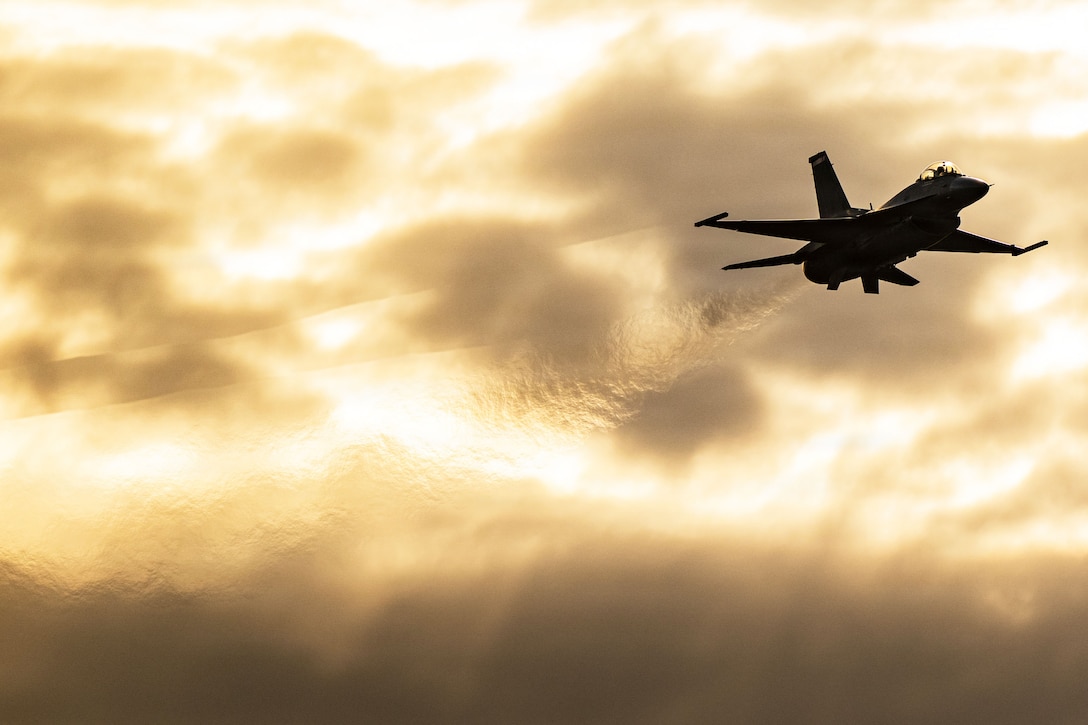 An Air Force aircraft in silhouette flies diagonally to the right in a golden, cloudy sky.