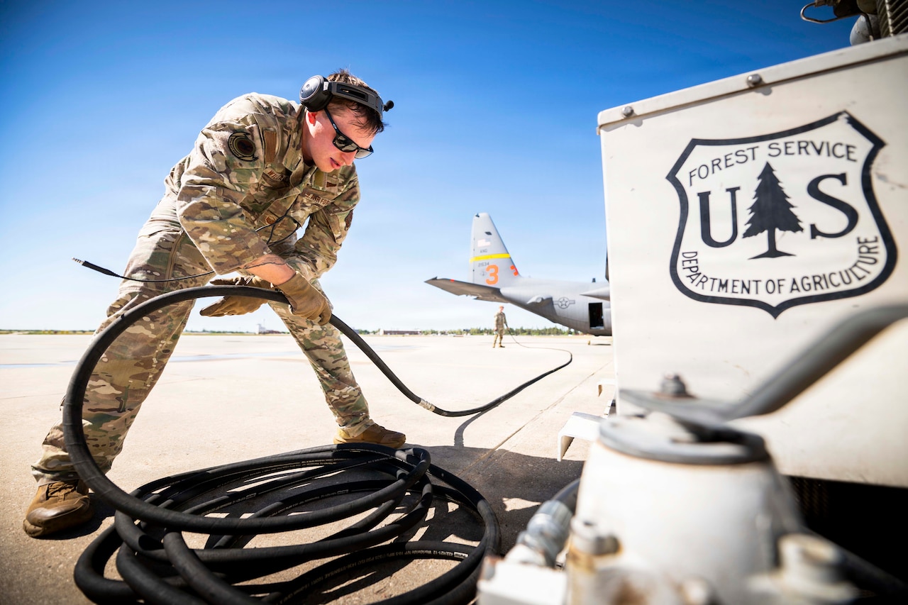 An airman unravels a black hose toward a fellow service member standing next to an aircraft on a tarmac during the day.