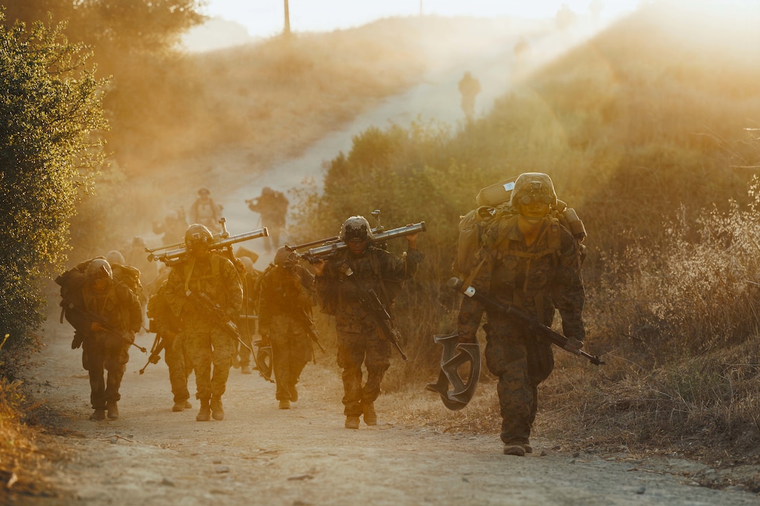 A group of Marines carrying rucksacks and other equipment hike behind a leader on a dirt path. Sunlight exposes a haze of dust.