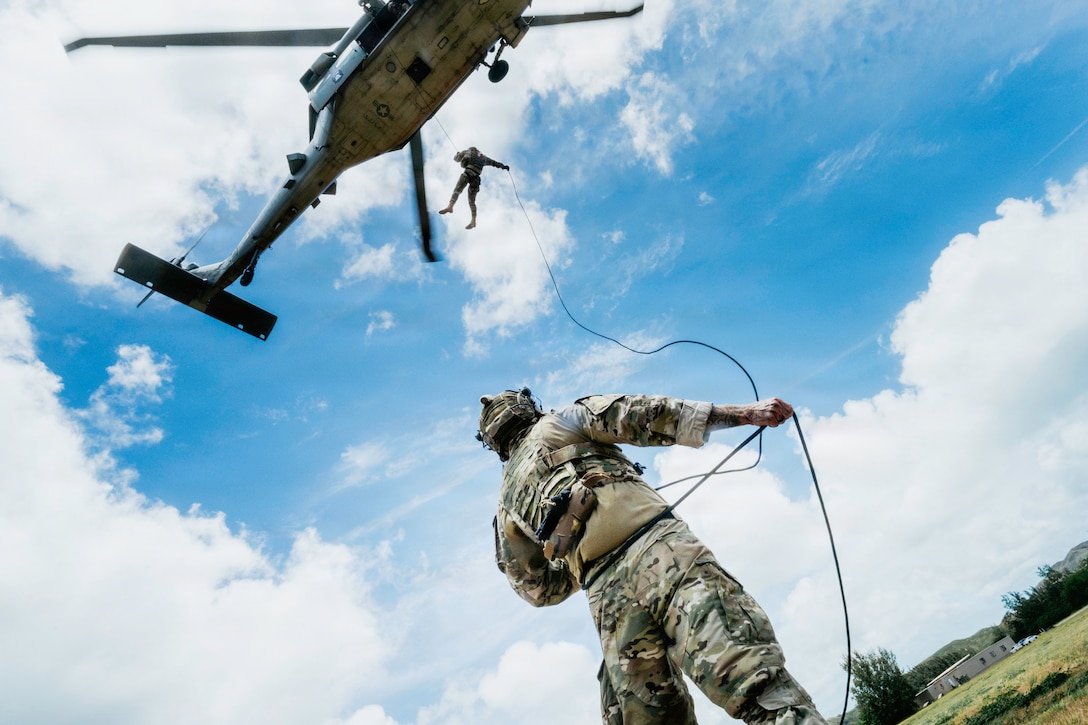 A service member holds onto a rope while hanging from a helicopter in the air, while another service member stands on the ground below holding the same rope.